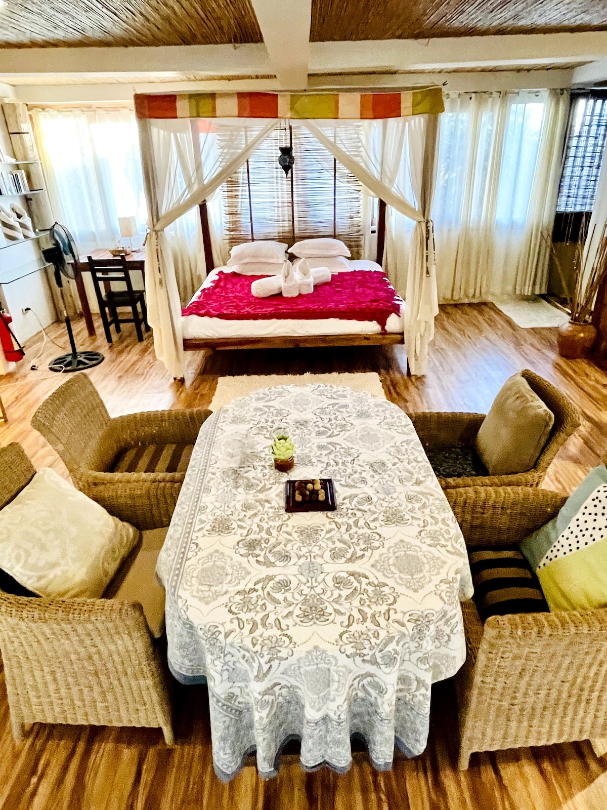The spacious living area features a king-size bed draped with curtains, complemented by a decorative red bedspread. A dining table for four with woven chairs is set nearby, enhancing the inviting atmosphere of the studio. Natural light filters through the sheer curtains, illuminating the wood flooring.