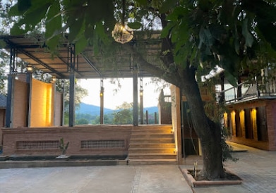 A tranquil outdoor area is framed by lush trees, featuring a stylish terraced structure. Soft lighting outlines the stairs leading to an elevated platform, with muted hues highlighting the natural surroundings and distant mountains in the background.