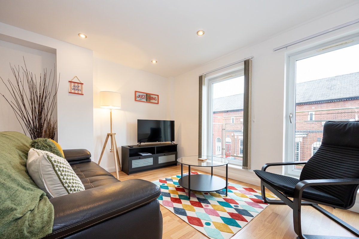 An open living area is highlighted by a sleek black sofa and an armchair, offering a relaxed seating arrangement. A television sits atop a low glass table, with vibrant rug patterns adding color to the hardwood floor. Large windows allow natural light to fill the space.