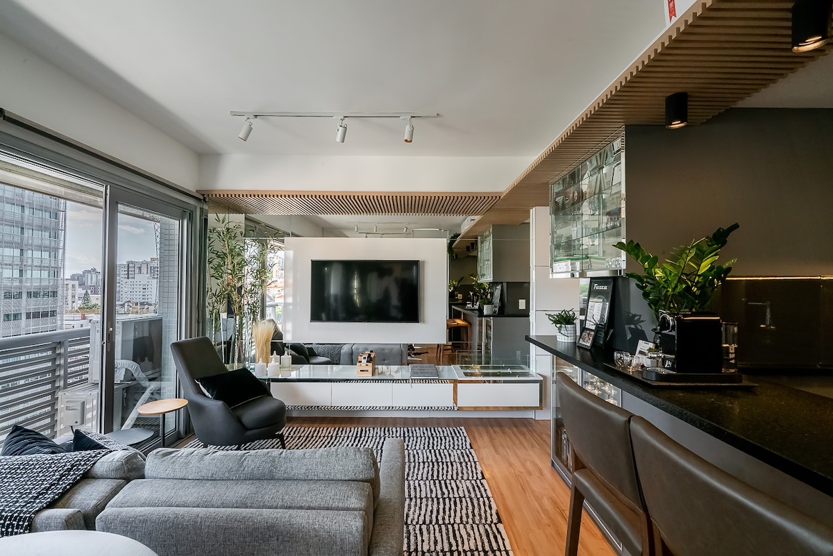 A modern living area is illuminated by natural light from large glass doors. A large television is mounted on the wall, alongside sleek cabinetry and decorative plants. Comfortable seating is arranged on a textured rug, creating a welcoming atmosphere for relaxation.