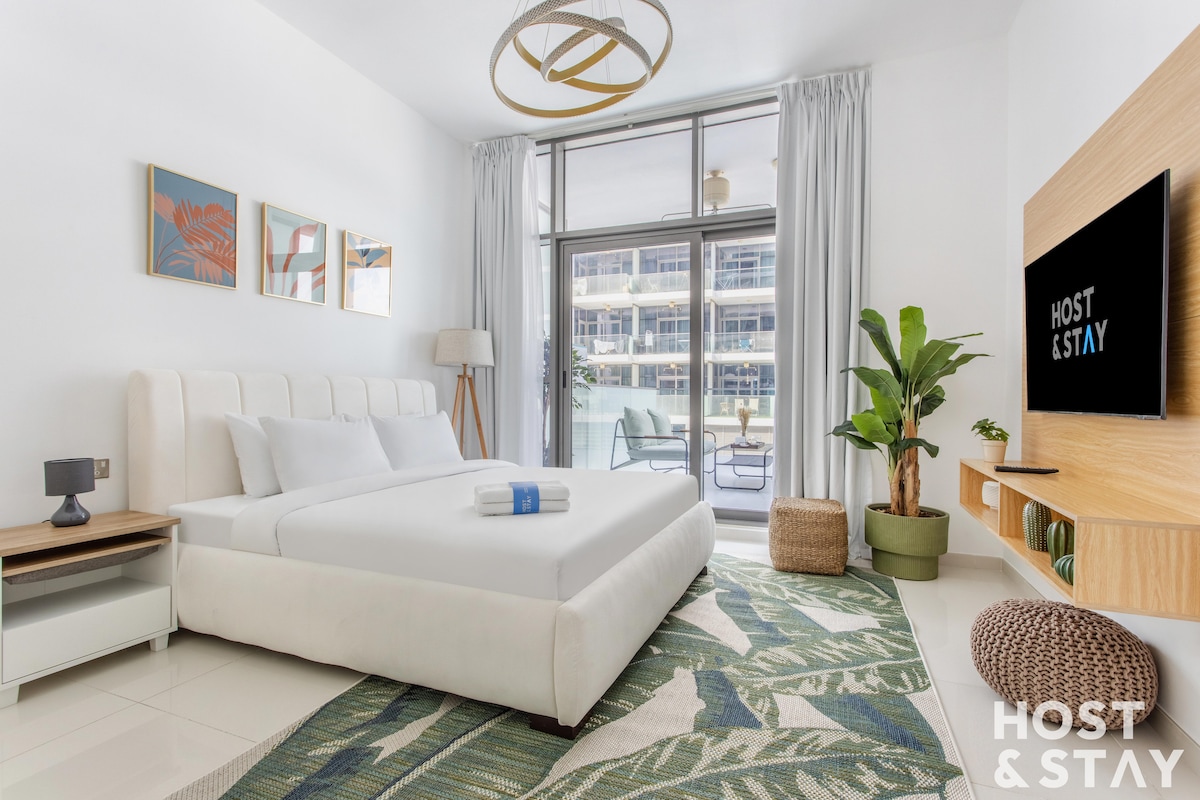 A modern studio is highlighted by a spacious bed with crisp white linens. A large television is mounted on the wall opposite, while a green area rug adds warmth to the room. Natural light fills the space through glass doors that open to a balcony.