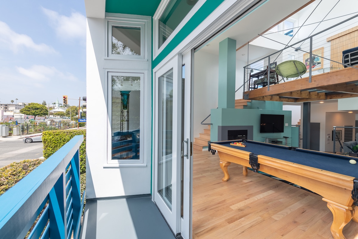 A modern balcony provides a view of the exterior and the nearby street, featuring large glass doors that open to the inside living area. A pool table is visible within the room, complemented by a fireplace and spacious layout with natural light flowing through the windows.