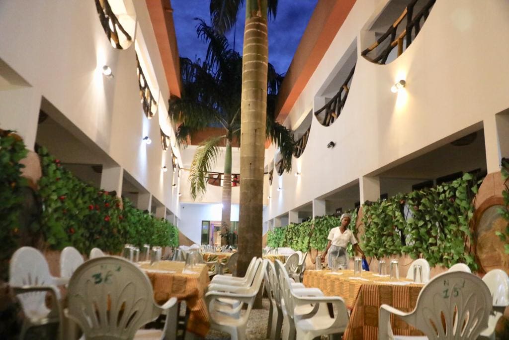 An open dining area is featured, with several white chairs arranged around tables covered in orange tablecloths. A tall palm tree stands in the center, surrounded by lush greenery along the walls and soft lighting that enhances the ambiance.
