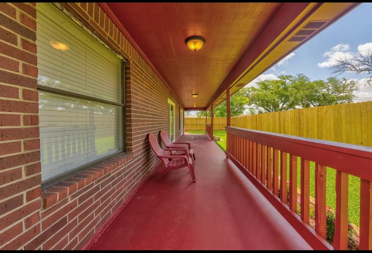 A covered porch features red flooring and brick walls, creating a cozy atmosphere. Two inviting rocking chairs are positioned to enjoy views of the green yard and wooden fence. Natural light filters through the windows, enhancing the cheerful outdoor space.