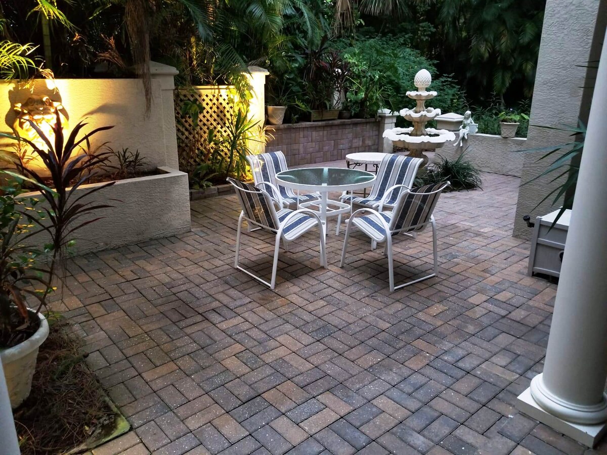 A serene outdoor patio area is presented, featuring a glass-top table surrounded by four striped chairs. Lush greenery borders the space, and a decorative fountain serves as a focal point, accompanied by soft lighting that enhances the tranquil ambiance.