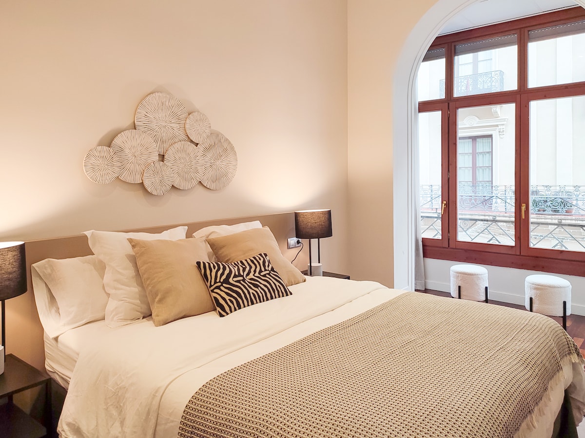 A serene bedroom space features a spacious bed adorned with neutral-toned pillows and a textured throw. Two bedside lamps provide gentle illumination, while a large window allows natural light to fill the room, offering views of the outside balcony.