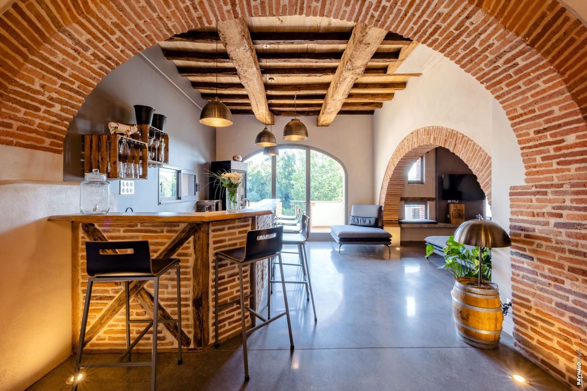 An inviting space features a rustic bar area with high stools, complemented by exposed wooden beams and stone archways. Large windows create an open ambiance, with natural light illuminating the seating area that includes a couch. A decorative plant and a barrel add to the charm.