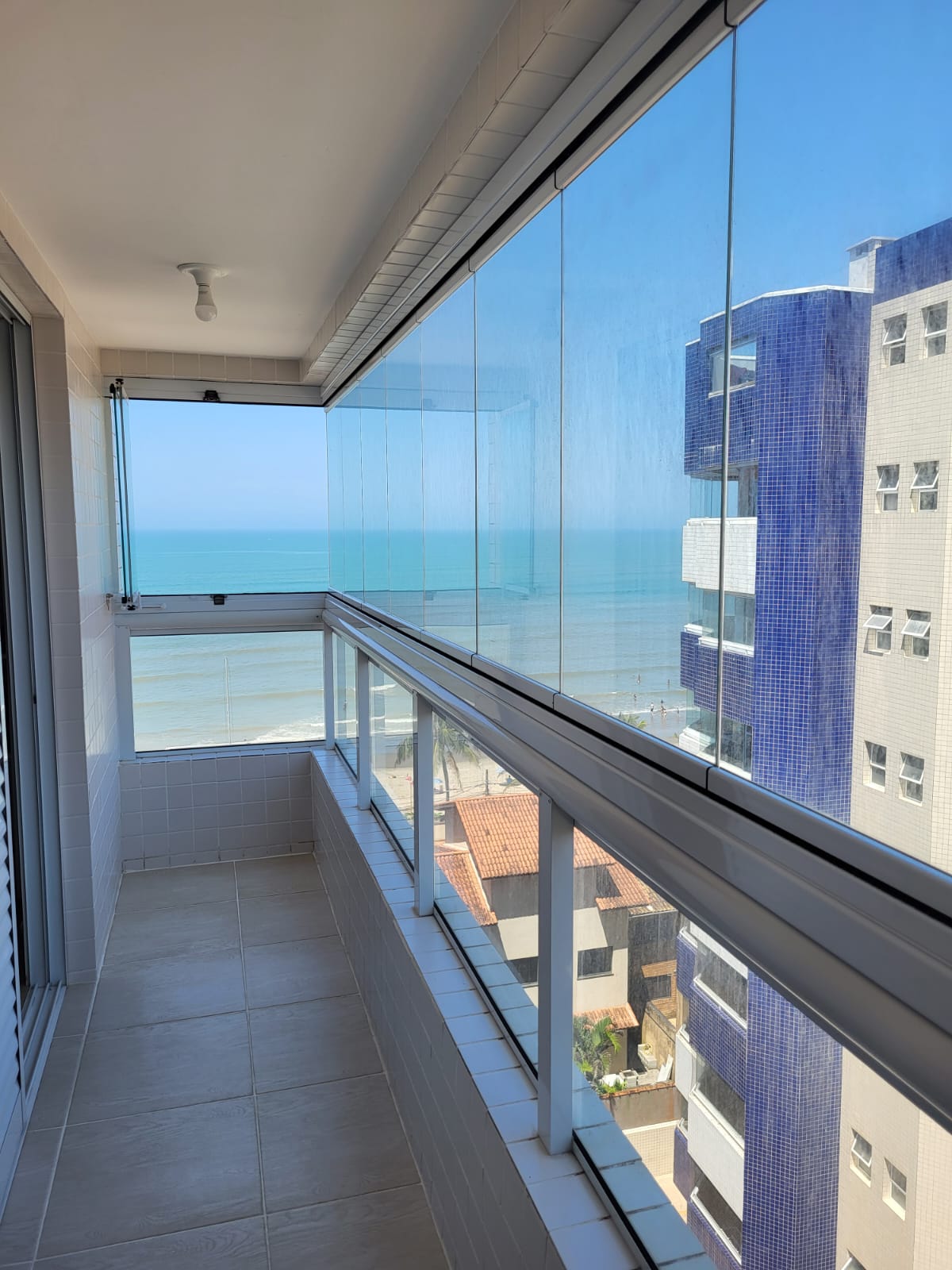 A balcony features transparent sliding glass panels offering unobstructed views of the ocean and nearby buildings. The calm blue waters reflect the clear sky, while the floor is tiled for easy maintenance. Natural light fills the space, creating a fresh and open ambiance.