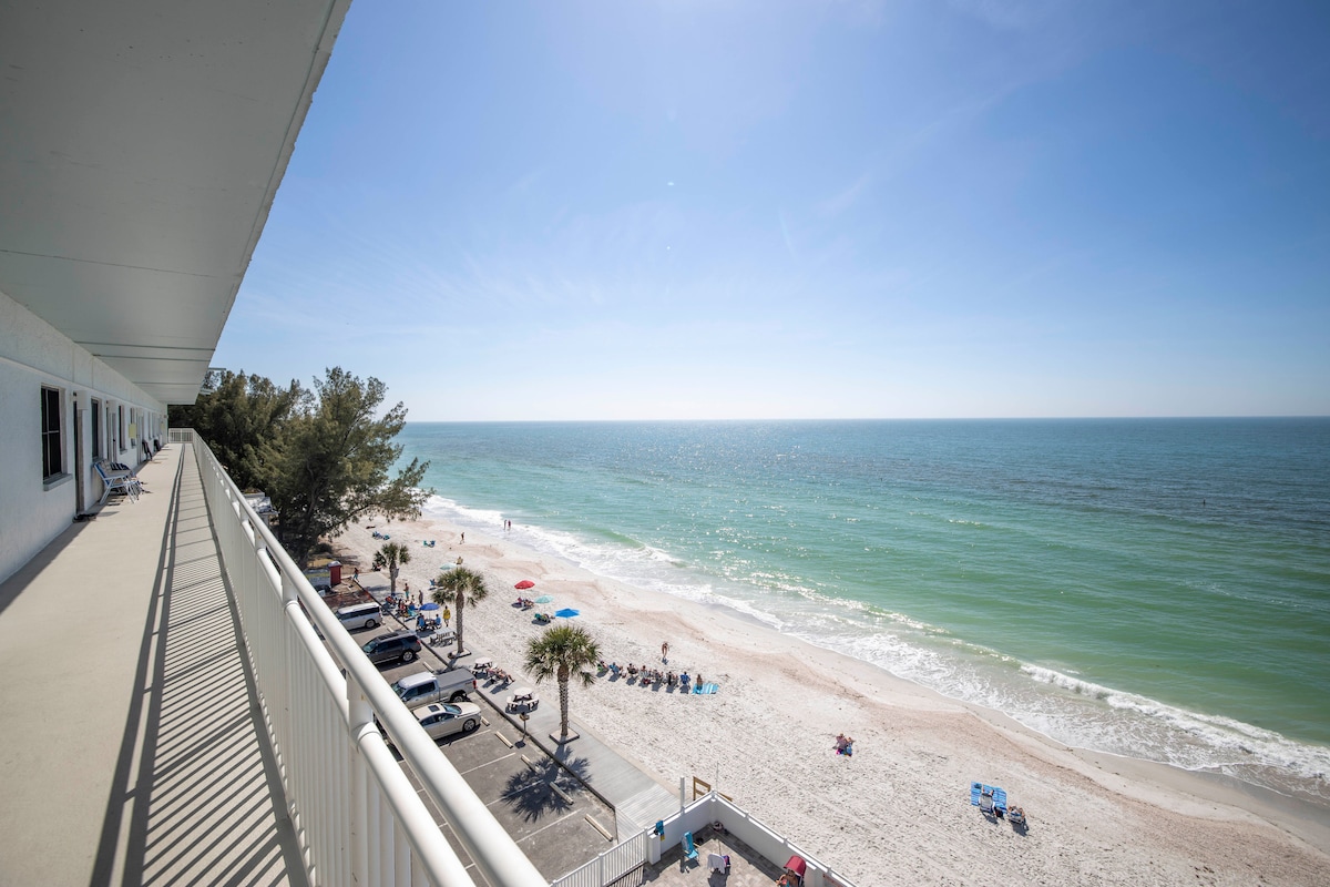 A spacious balcony offers a direct view of the sandy beach and tranquil Gulf waters. Sun chairs can be seen arranged along the shore, while a few palm trees provide shade. The bright blue sky enhances the serene coastal atmosphere.
