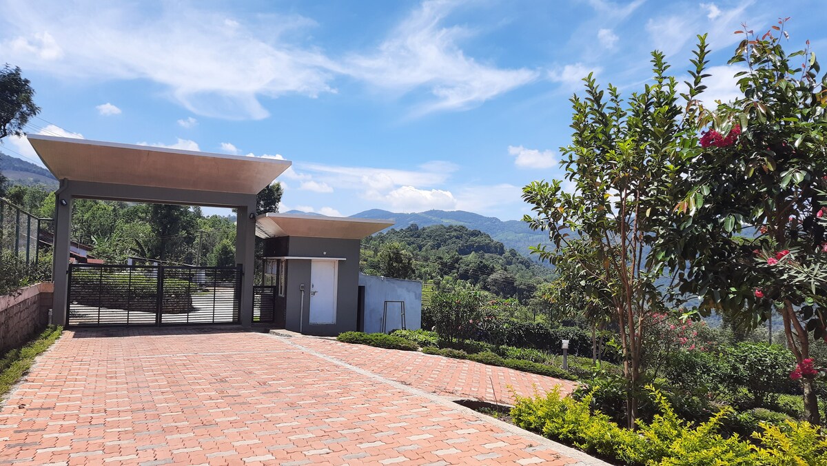 The entrance features a spacious driveway paved with red bricks, flanked by manicured greenery and blooming flowers. A modern gate opens to reveal scenic hills and a clear sky, inviting visitors into the tranquil setting of the tea estate.