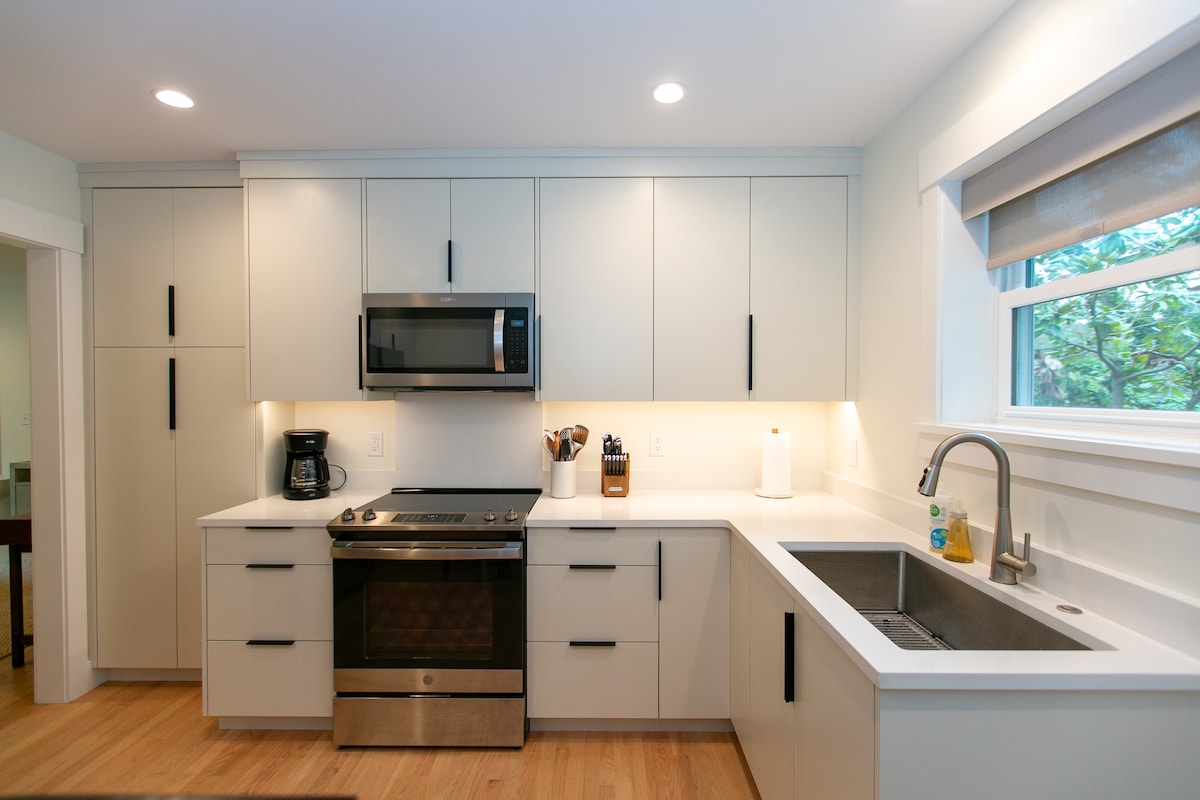 A modern kitchen features sleek cabinetry and stainless steel appliances. A built-in microwave and coffee maker are positioned on the countertop. A large window allows natural light to illuminate the space, while the sink and stovetop are integrated into the minimalist design.