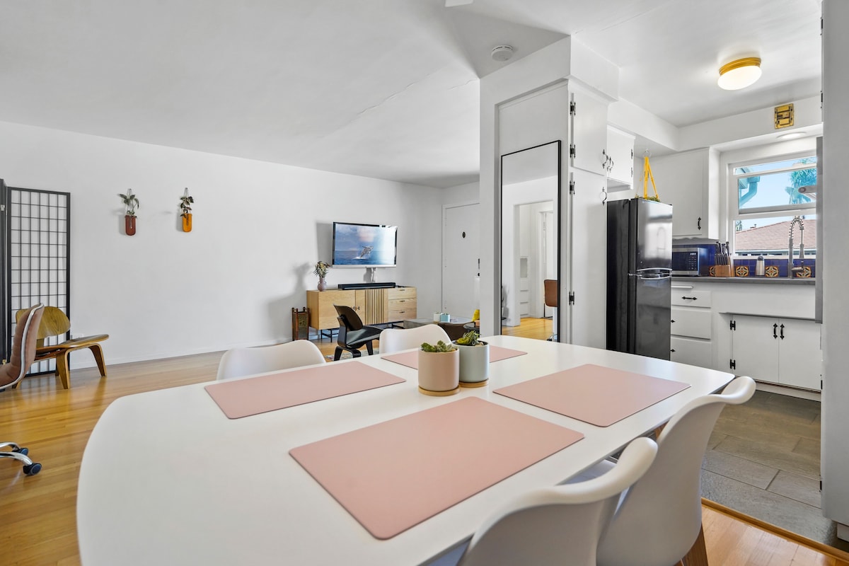 A bright living space features a spacious dining table surrounded by modern white chairs. A sleek entertainment unit holds a flat-screen TV, while a kitchen area is visible in the background with black appliances and a window inviting natural light.
