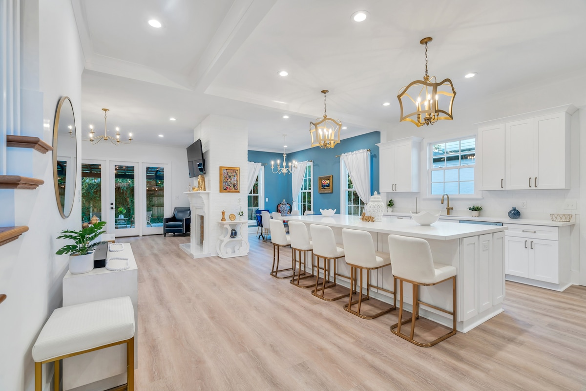 A bright and airy open living space is highlighted by elegant lighting fixtures and wooden flooring. The kitchen features a large island with seating for six, while large windows provide ample natural light. A cozy fireplace is seen in the background, enhancing the ambiance.