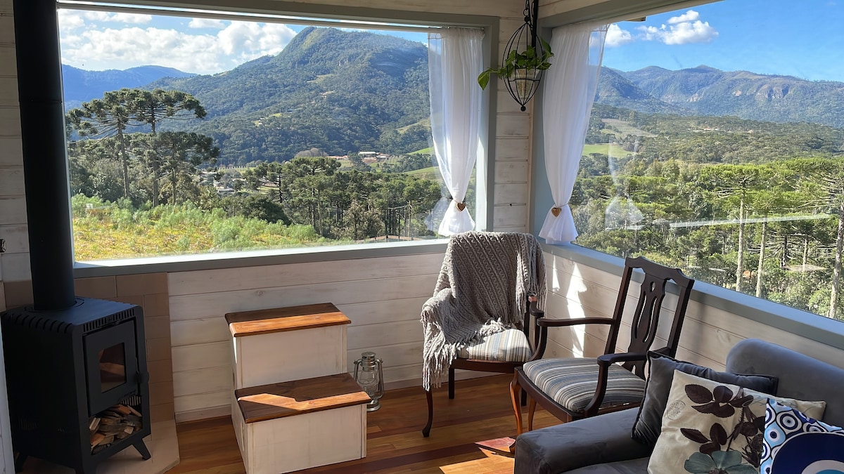 A cozy living area features large windows that provide expansive views of the surrounding mountains. A comfortable chair and blanket are positioned beside a wood stove. A small set of wooden steps complements the arrangement, creating an inviting corner for relaxation.