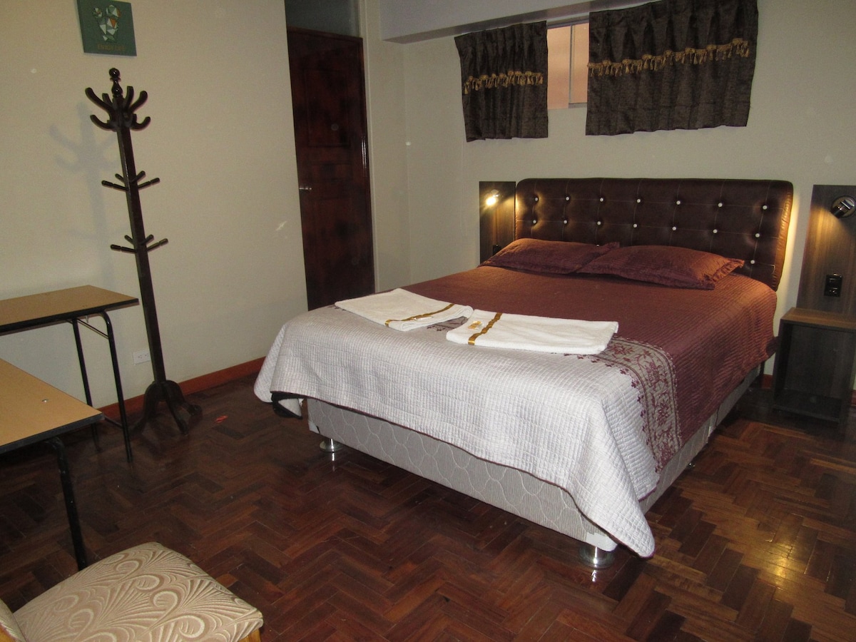 A spacious bedroom features a queen-size bed with a dark headboard and crisp linens. A wooden table with a chair and a decorative coat rack are present in the room. Light filters through the patterned curtains, enhancing the warm tones of the wooden flooring.