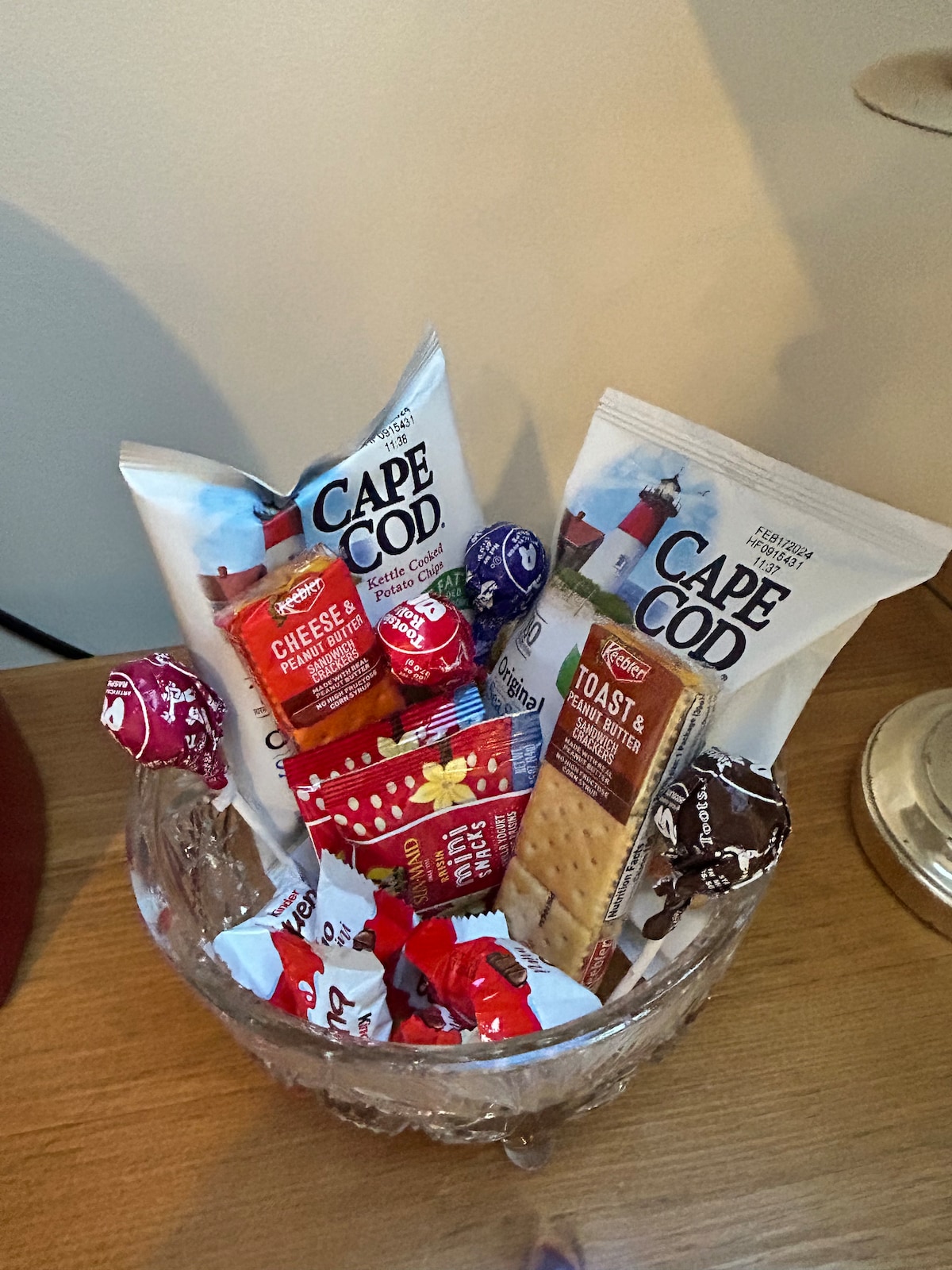An assortment of snacks is displayed in a decorative glass bowl. The selection includes bags of Cape Cod chips, cheese crisps, and various candies wrapped in colorful packaging, offering a variety of savory and sweet options for guests.
