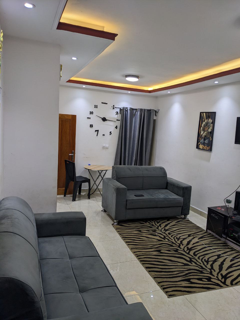 The living area features two comfortable gray sofas positioned on a soft area rug with a zebra pattern. A small dining table is present, accompanied by a black chair. A stylish clock adorns the wall, with modern lighting highlighting the space.