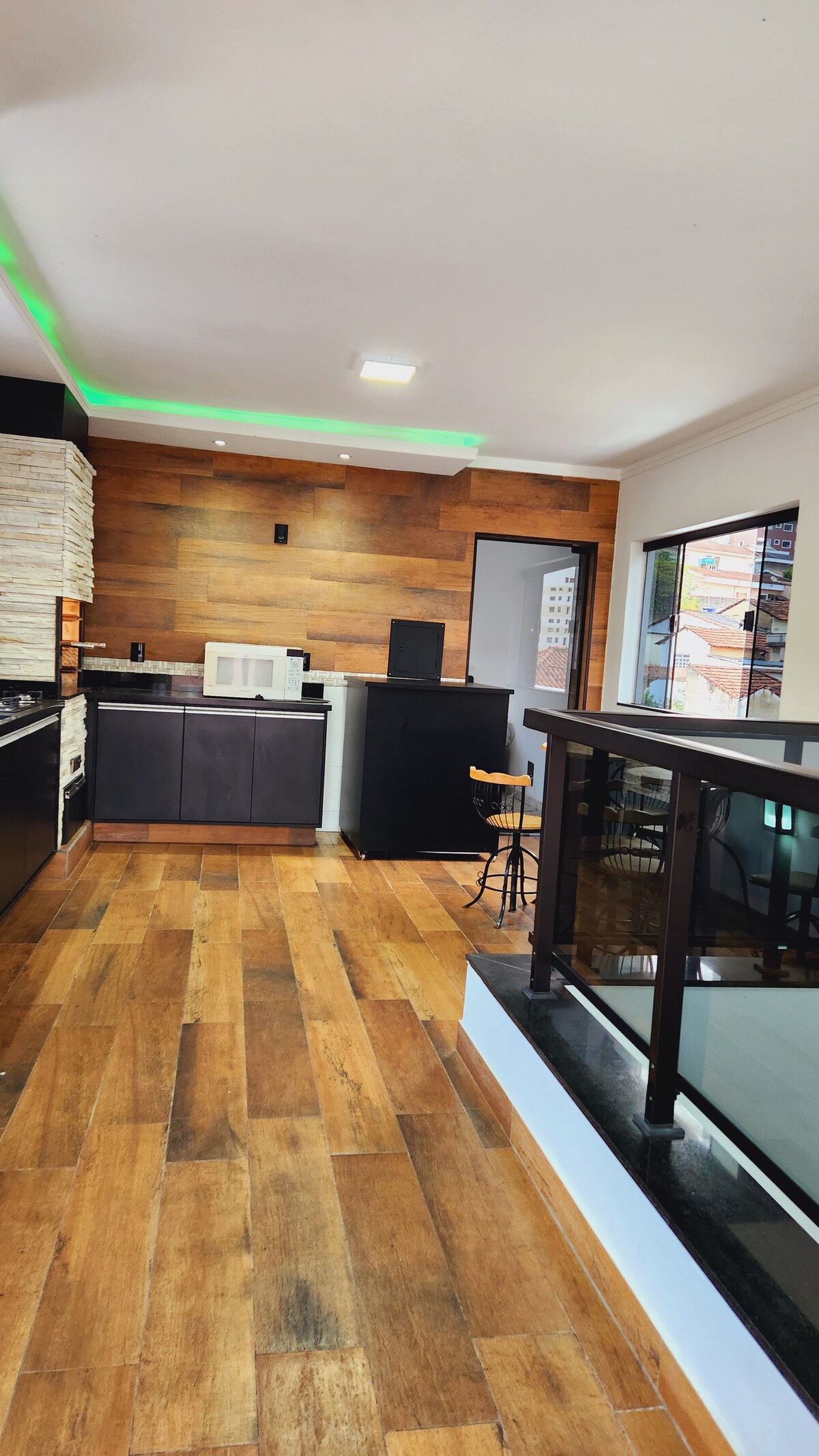 A modern kitchen area is shown, featuring sleek black cabinets and a light-colored countertop. Warm wooden accents create a cozy ambiance. Natural light enters through a window, highlighting the open layout and a small bar area with a stool. Flooring is composed of wooden planks.