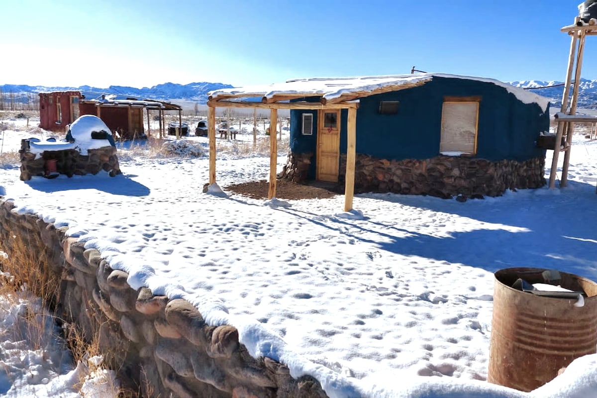 A cozy cabin is nestled among a blanket of snow, featuring a stone exterior and a wooden porch. The clear blue sky provides a bright backdrop, while mountains are faintly visible in the distance. Nearby, a stone structure and metal barrel are also present.