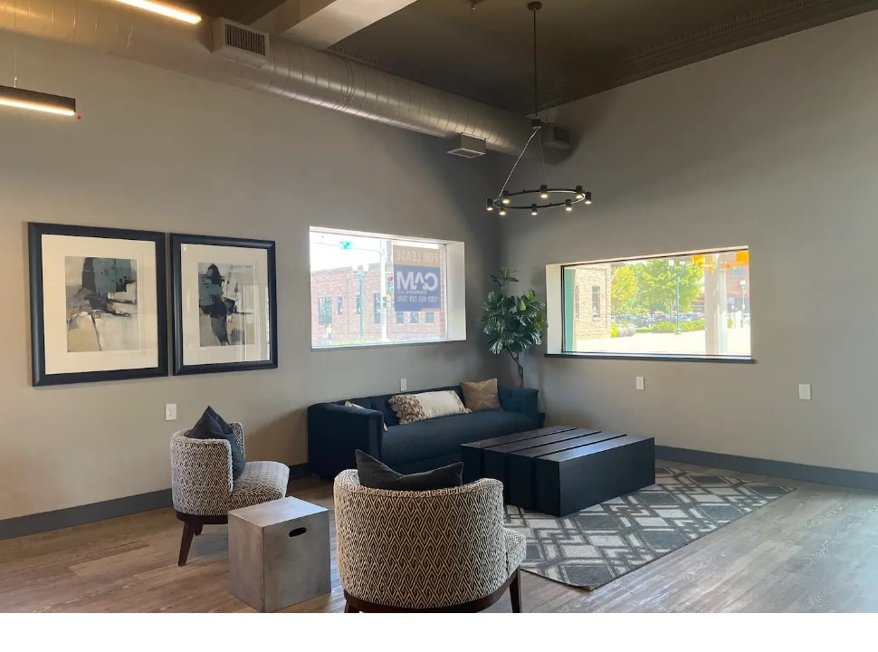 A shared lobby area is featured, showcasing a modern seating arrangement with a black sofa and two patterned armchairs. Large windows allow natural light to illuminate the space, complemented by minimalist wall art and a geometric area rug.