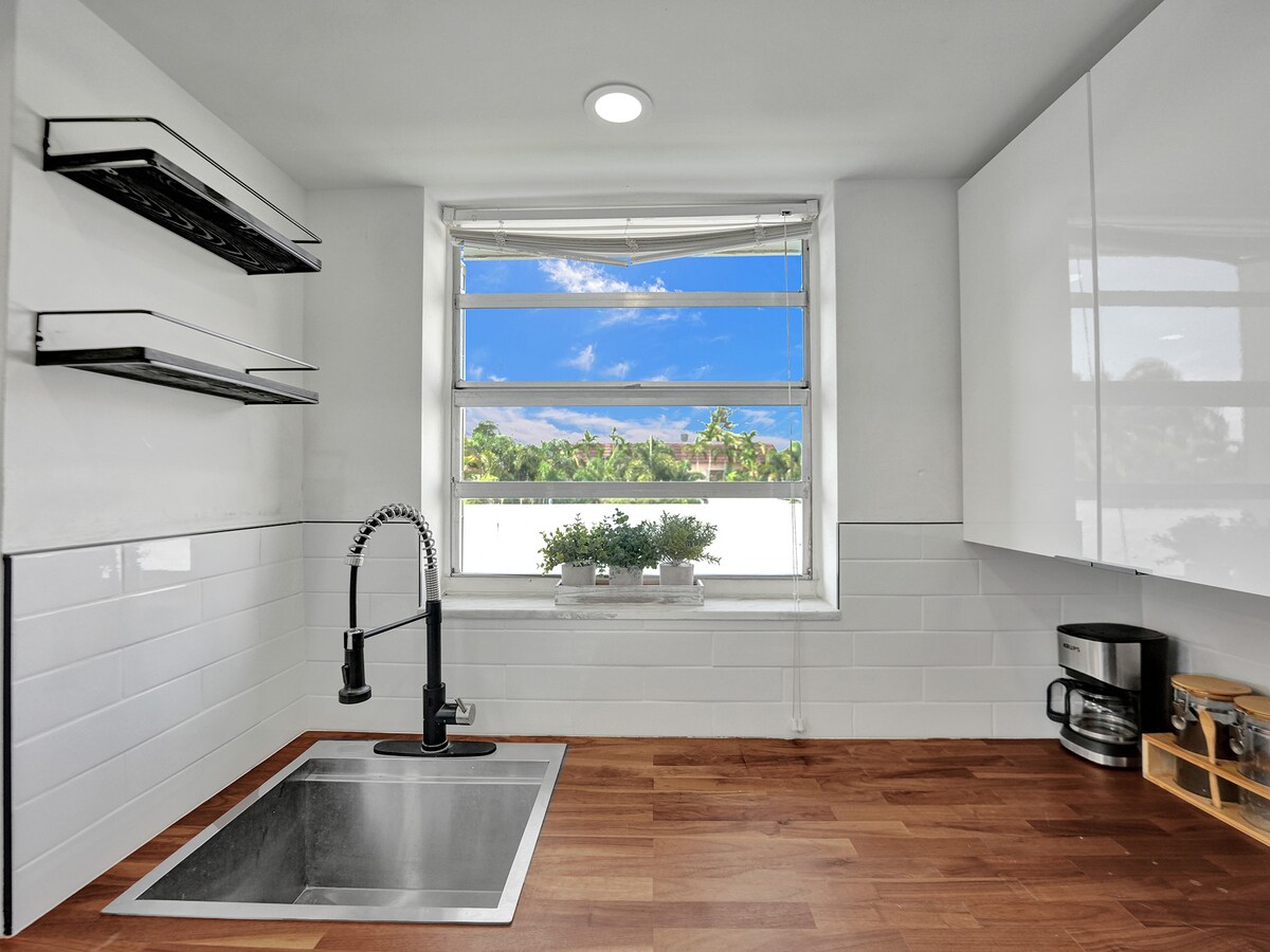 The kitchen features white cabinetry and a sleek, modern countertop. A window offers a view of the outdoors, framed by natural light. A coffee maker and a small storage rack can be seen, enhancing the practicality of the space.