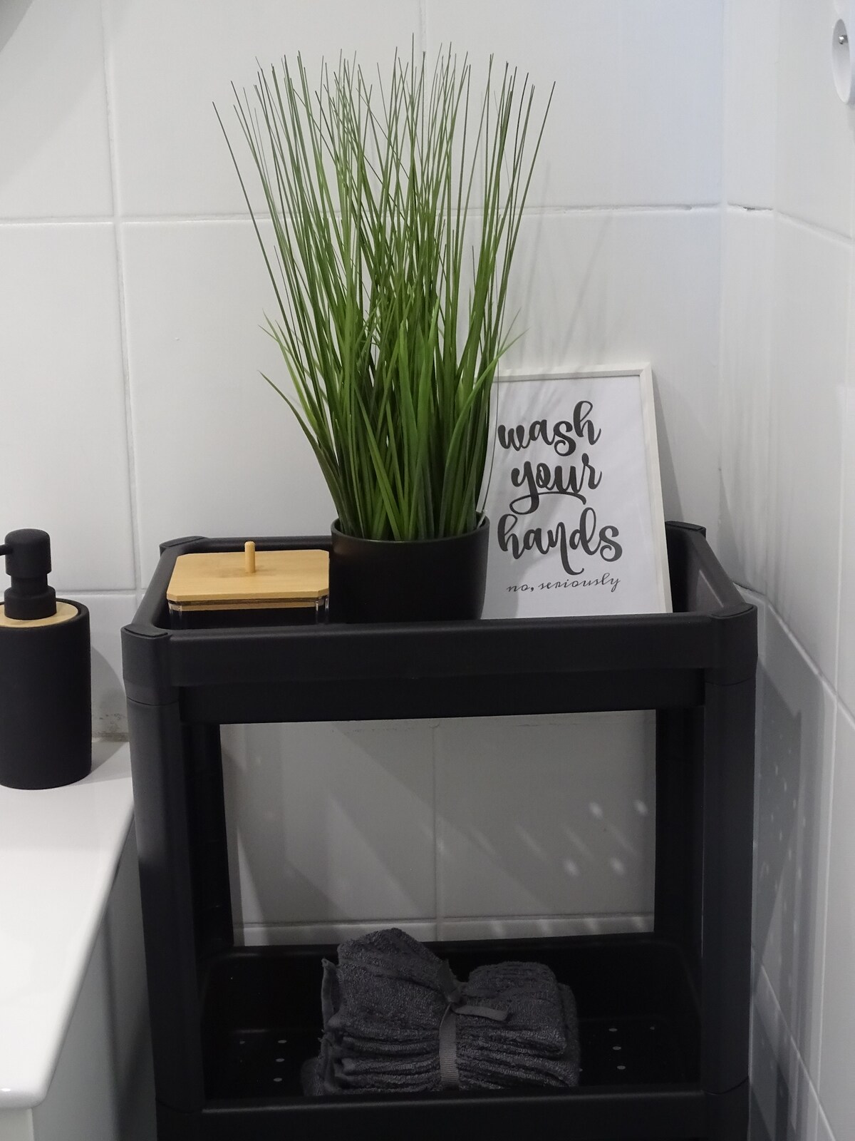A bathroom arrangement features a black shelving unit holding neatly folded dark towels. A small wooden tray and a soap dispenser are positioned beside a potted plant, while a sign with the phrase 'wash your hands' adds a touch of decor.