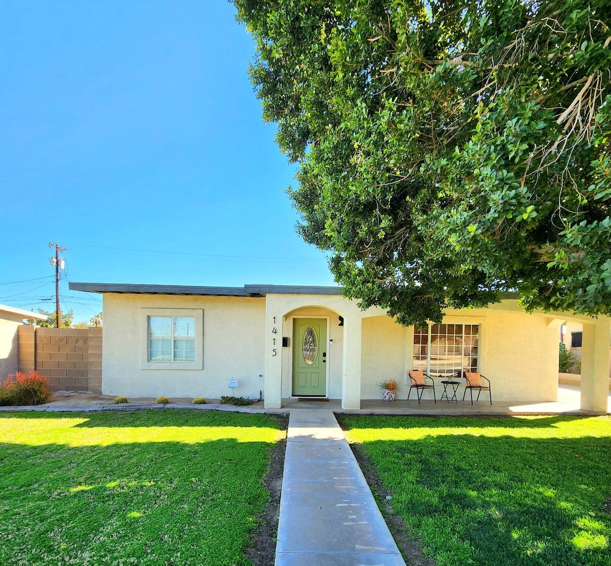A single-story home is framed by a well-maintained lawn, featuring a welcoming front porch with two chairs and a round table. A large tree provides shade, and the entrance door, flanked by decorative windows, adds charm to the exterior.