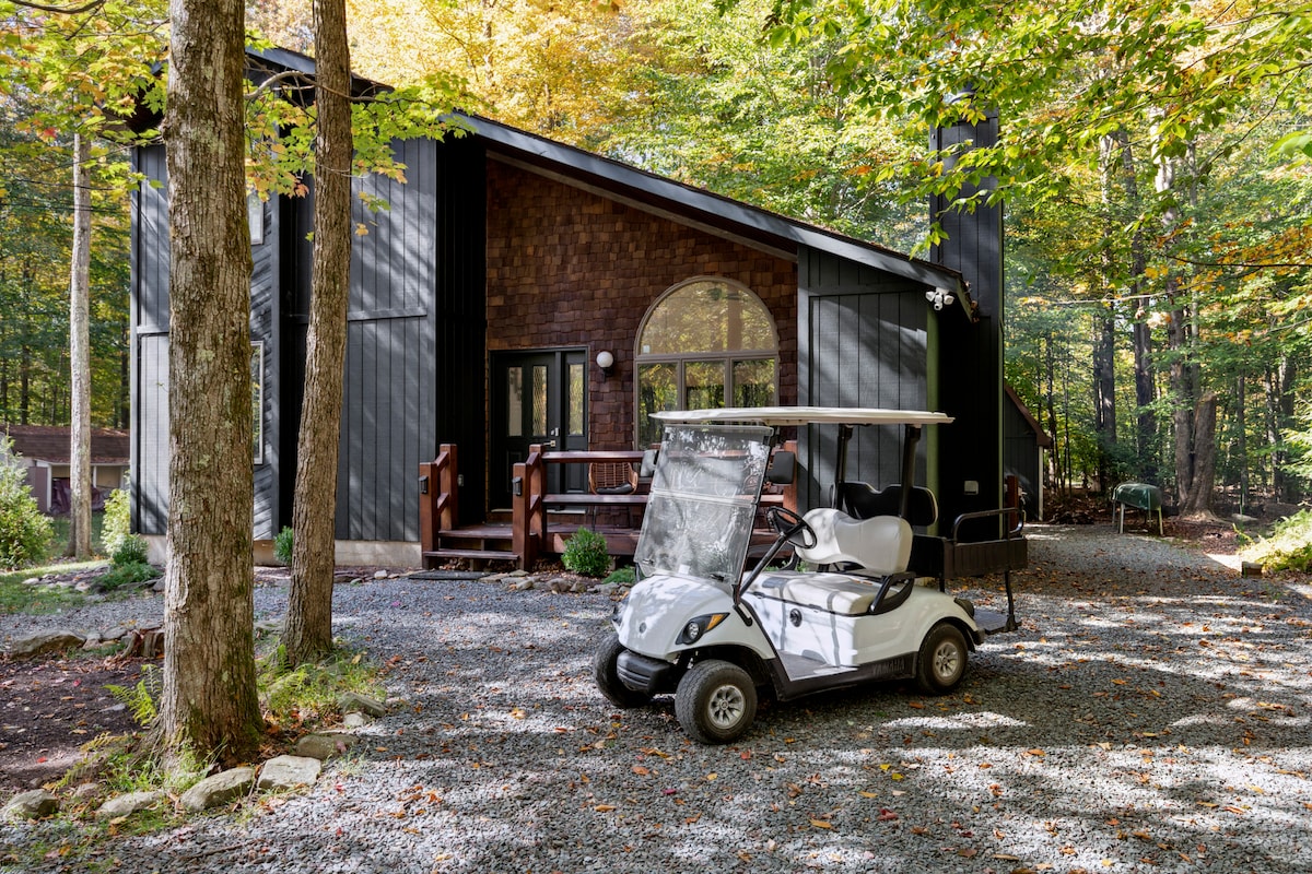 A golf cart is parked in front of a modern-style cabin, surrounded by lush trees showcasing autumn foliage. The exterior features a blend of dark wood and metal accents, with large arched windows offering a glimpse of the inviting interior.