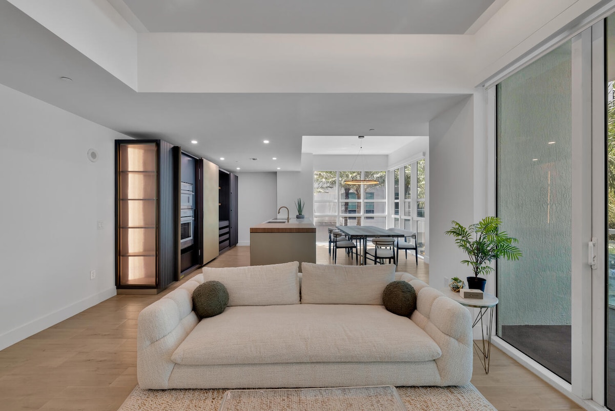 A contemporary living area features a light-colored sofa with large, round cushions. A stylish area rug complements the space. Natural light floods in from expansive glass doors leading to a balcony. A dining area and modern kitchen are visible in the background.