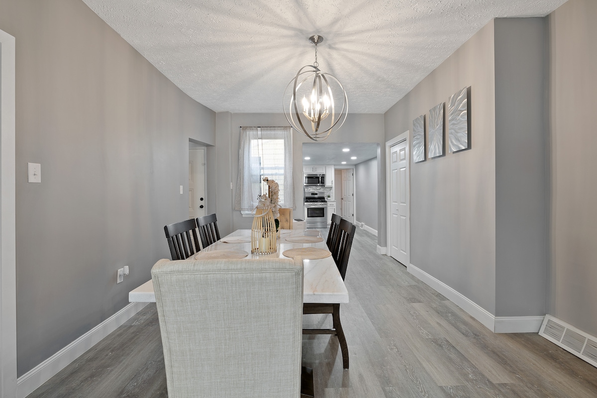 The dining area features a large, rectangular table set for meals, surrounded by dark wooden chairs and a light-colored upholstered chair. A modern light fixture hangs above, illuminating the space. Neutral grey walls and light flooring enhance the room's contemporary design, promoting a welcoming atmosphere.