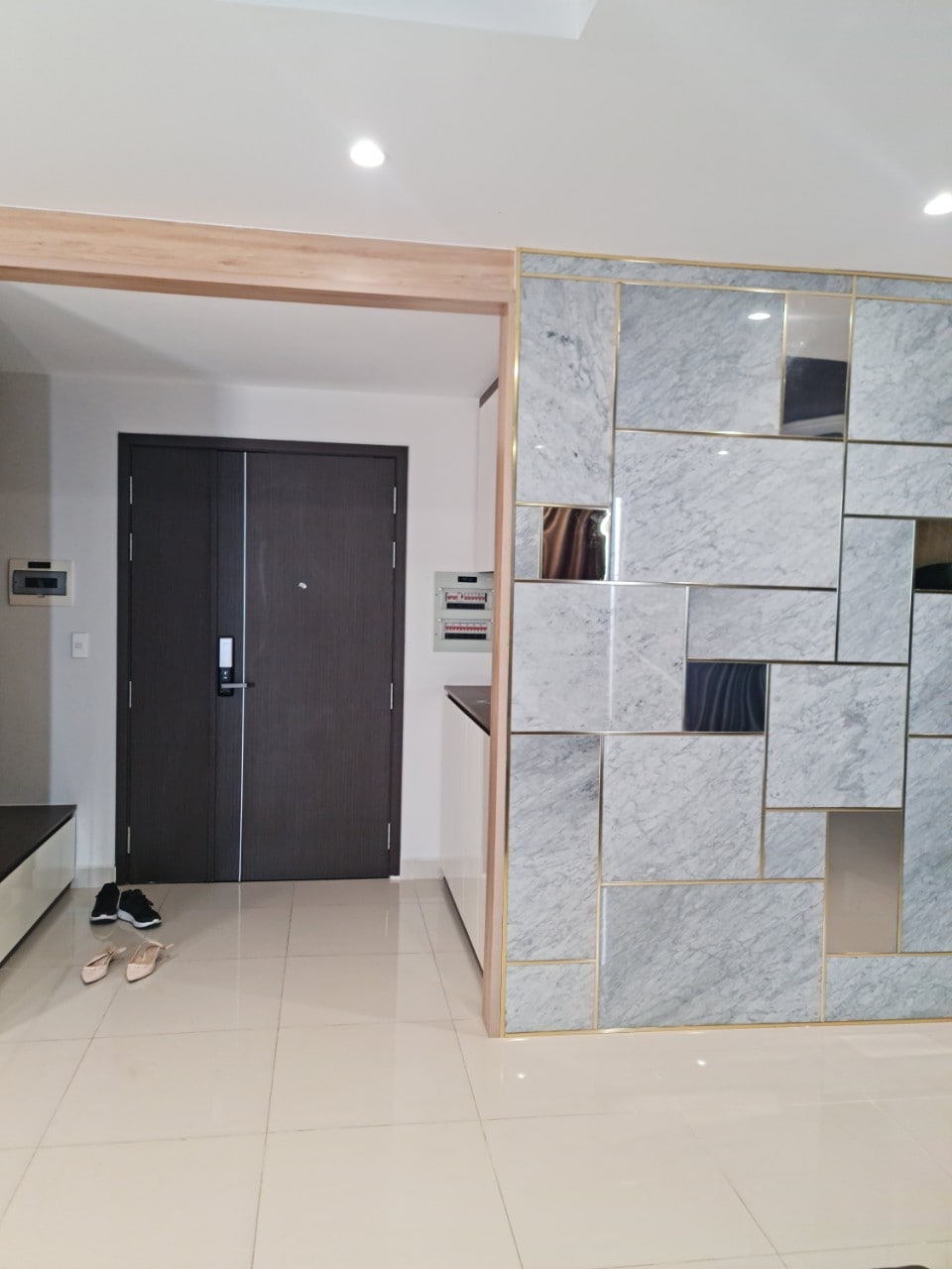 An entrance area features a modern design with a textured wall of gray marble squares outlined in gold. A stylish bench is positioned against one wall, with a pair of shoes placed neatly in front of the entrance door.