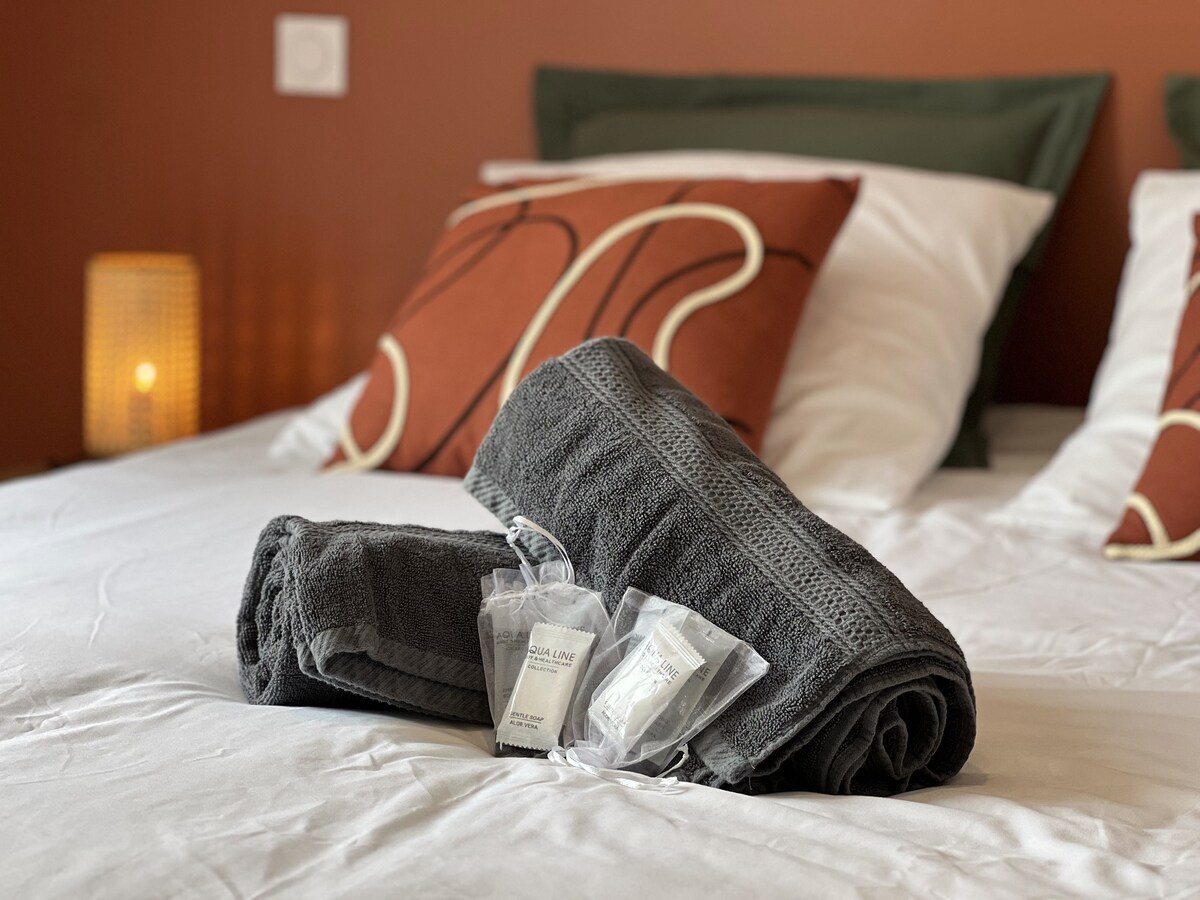A neatly made bed is presented with two pillows adorned with decorative cases. Soft, dark towels are arranged with complimentary toiletries nearby. A softly glowing lamp adds a warm touch to the cozy bedroom setting.