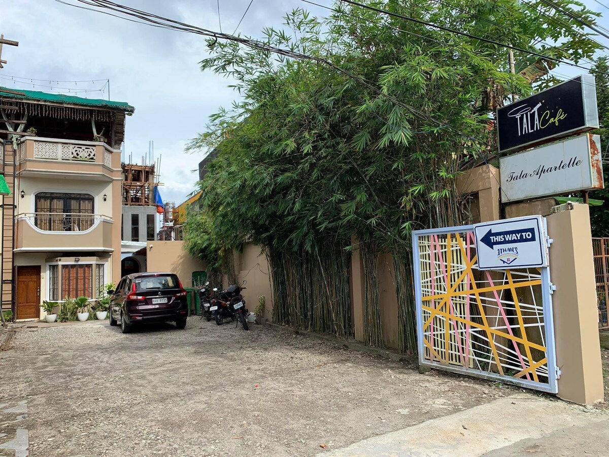 An entrance view of Tala Apartelle is depicted, showcasing a gravel parking area accommodating vehicles. A modern sign displaying 'Tala Apartelle' is positioned on the right, while bamboo creates natural greenery along the left side, enhancing the welcoming entrance to the property.