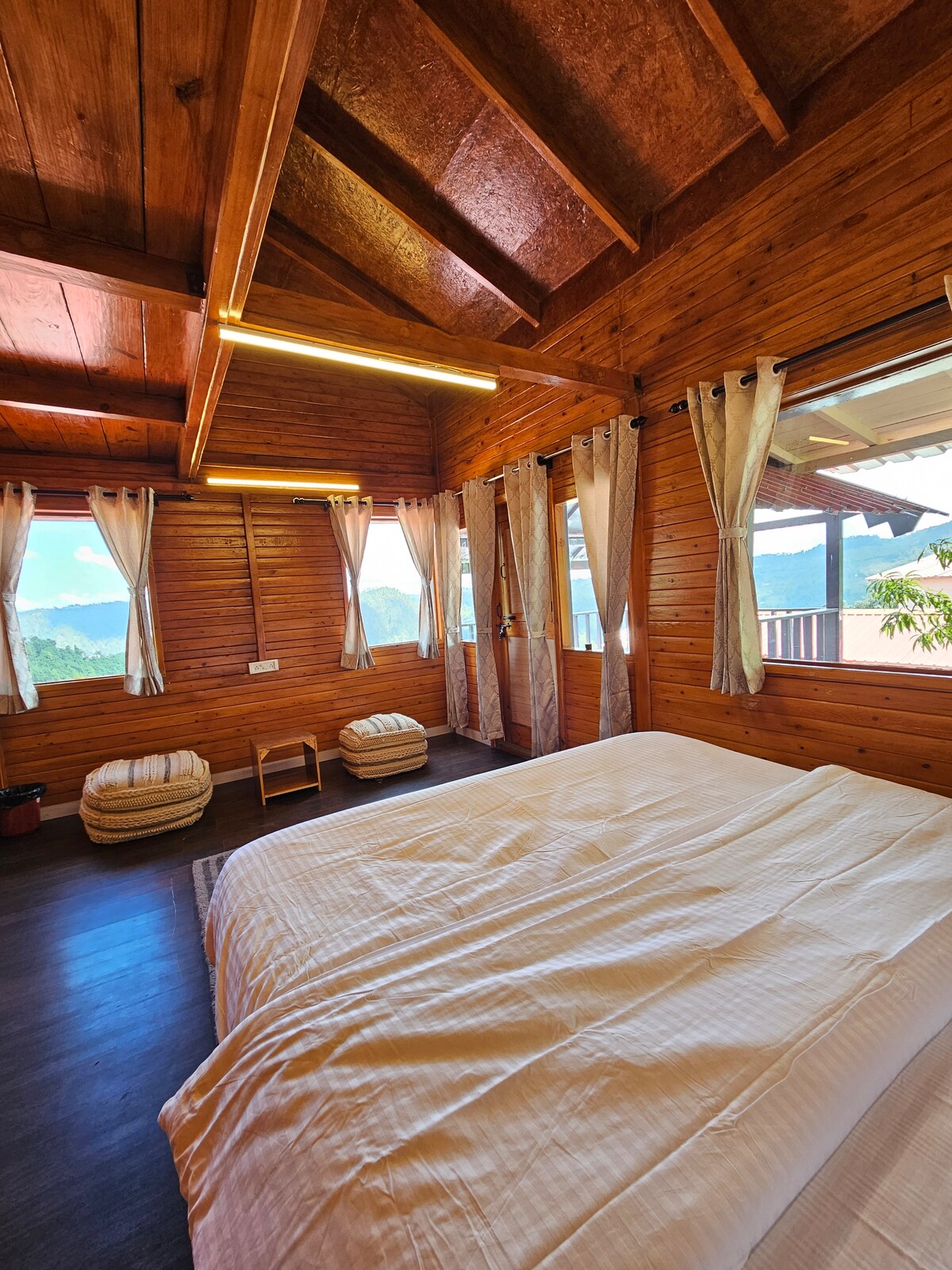 A wooden-walled room features a spacious bed with crisp white linens. Large windows are adorned with light-colored curtains, allowing natural light to fill the space. Two cozy floor cushions are placed nearby, providing additional seating options. Scenic views of greenery can be seen through the windows.