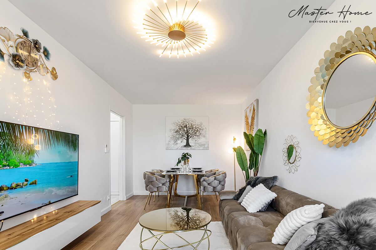 The spacious living area features a large, wall-mounted television and a comfortable sofa adorned with soft cushions. A modern gold chandelier illuminates the room, while a round coffee table sits at the center. A dining area with stylish chairs and a decorative wall art piece enhances the ambiance.