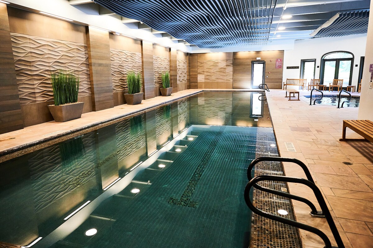 An indoor pool area features a tranquil swimming pool, bordered by elegant wooden lounge chairs. Contemporary stone walls and planters with greenery create a calm atmosphere. Soft lighting reflects off the water's surface, enhancing the serene environment.