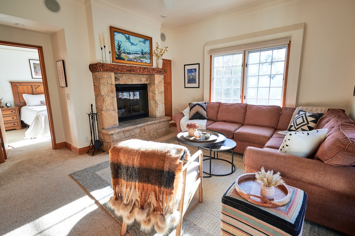 A cozy living area features a large sectional sofa adorned with various cushions and a soft throw. A stone fireplace is positioned against the wall, providing warmth. Natural light enters through large windows, illuminating the space, while a circular coffee table and a decorative side table complete the setup.