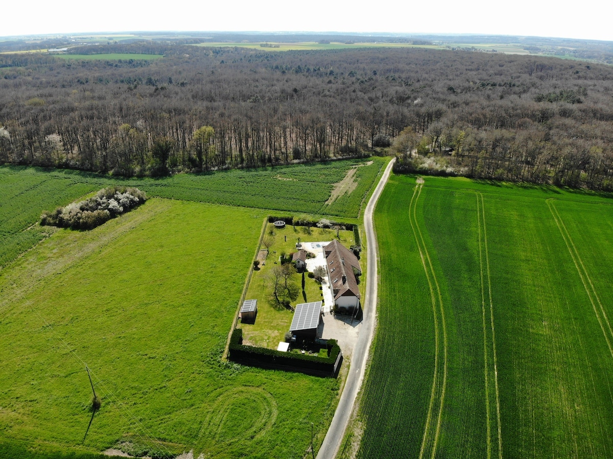 An aerial view captures a charming house surrounded by expansive green fields and a garden. The home is nestled within a spacious outdoor area, featuring a variety of landscaping, including trees and open space. The surrounding countryside is visible, highlighting the tranquil setting.