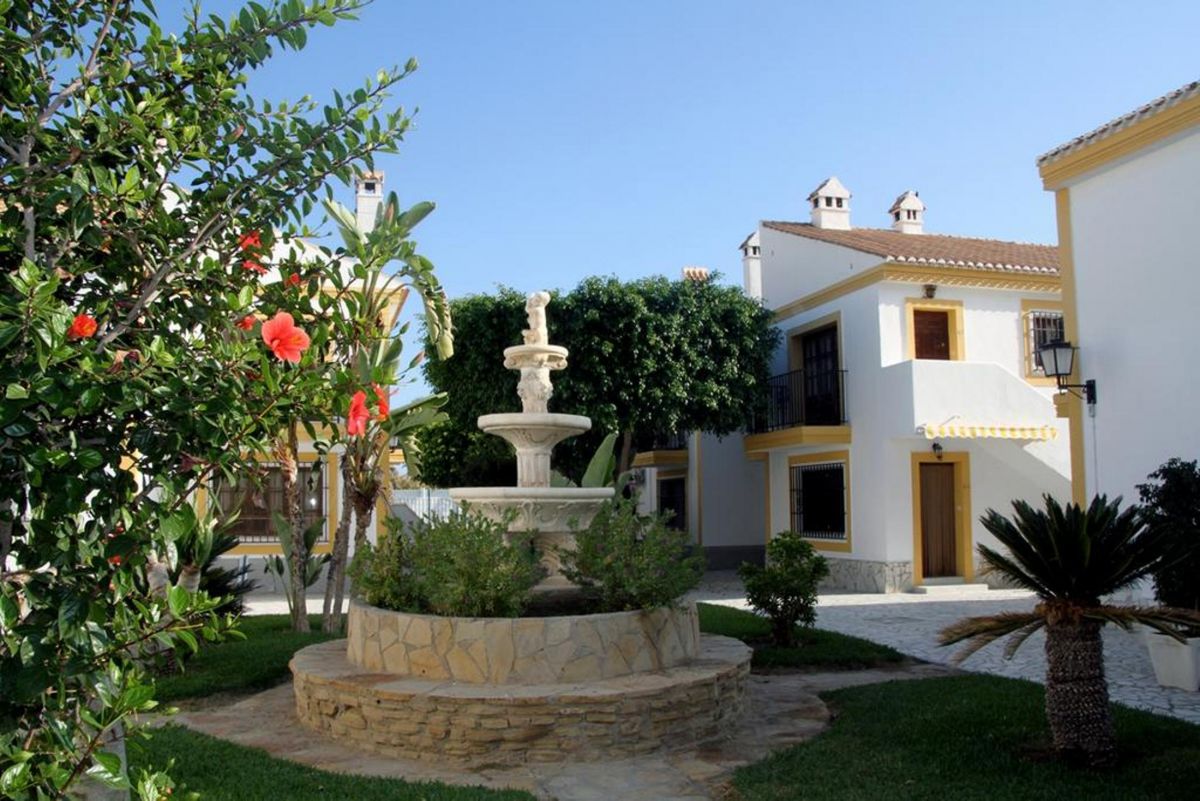 A charming communal garden is featured, showcasing a stone fountain surrounded by lush greenery and colorful flowers. The well-maintained area includes paths leading to residential buildings, creating a serene and inviting atmosphere. Bright blue skies complement the scene.