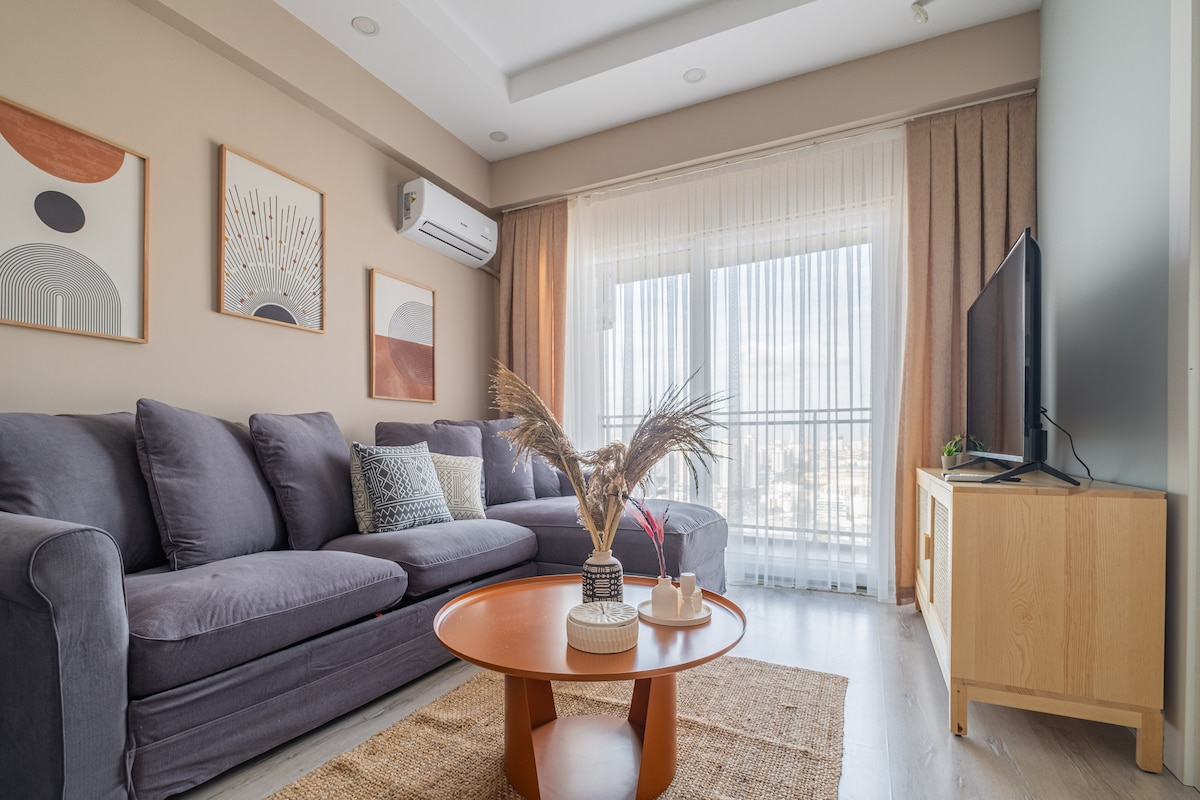 A comfortable living area is presented with a gray sofa arranged around a circular wooden coffee table. Decorated with framed artwork, the space offers large windows dressed with sheer curtains, allowing natural light to illuminate the room. A flat-screen television is positioned on a light wooden media unit.