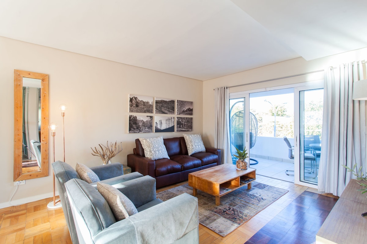 A spacious living area features a combination of comfortable seating, including a leather sofa and fabric chairs. Natural light filters through large glass doors that lead to an outdoor area. Decorative wall art adds character, while a wooden coffee table anchors the space.