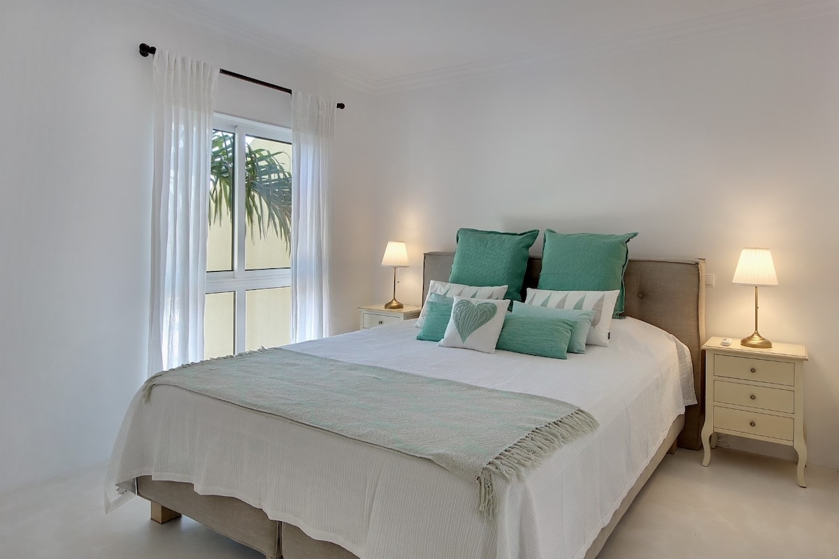 Chambre principale avec grand lit, fenêtre pleine longueur avec stores/volets pour une obscurité totale, armoire ouverte, commode, climatisation (chaud/froid) et salle de bains privative avec baignoire complète, douche (sur baignoire), toilettes et lavabo avec vasque et miroir