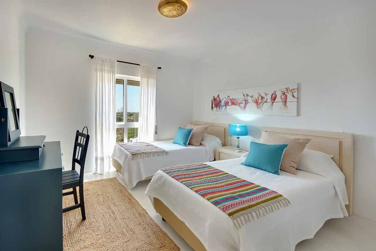 Troisième chambre avec deux lits séparés, fenêtre pleine longueur avec stores/volets pour une obscurité totale, armoire ouverte, dressing, chaise et miroir, climatisation (chaud/froid) et accès à la salle de douche avec douche à l'italienne, toilettes et lavabo avec vasque et miroir