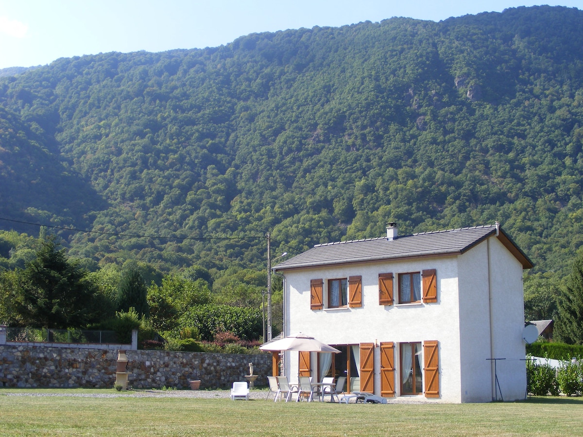 A two-story house is set against a backdrop of lush mountains. The exterior features light-colored walls and wooden shutters. A patio area is visible, furnished with sun loungers and an umbrella, all surrounded by a grassy expanse and trees.