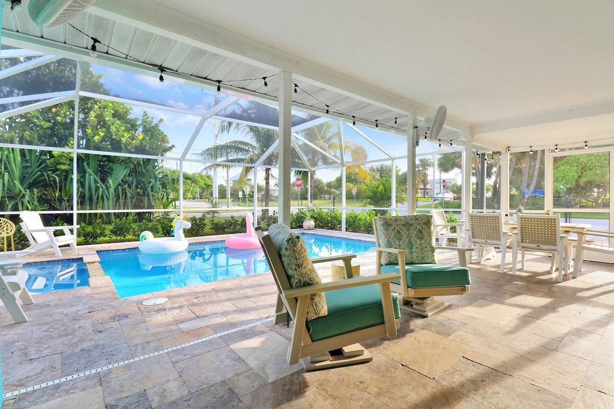 A spacious outdoor living area features a heated pool, surrounded by palm trees and tropical plants. Comfortable rocking chairs are positioned nearby, offering a relaxing view. Brightly colored inflatable pool floats add a playful touch to the inviting space.