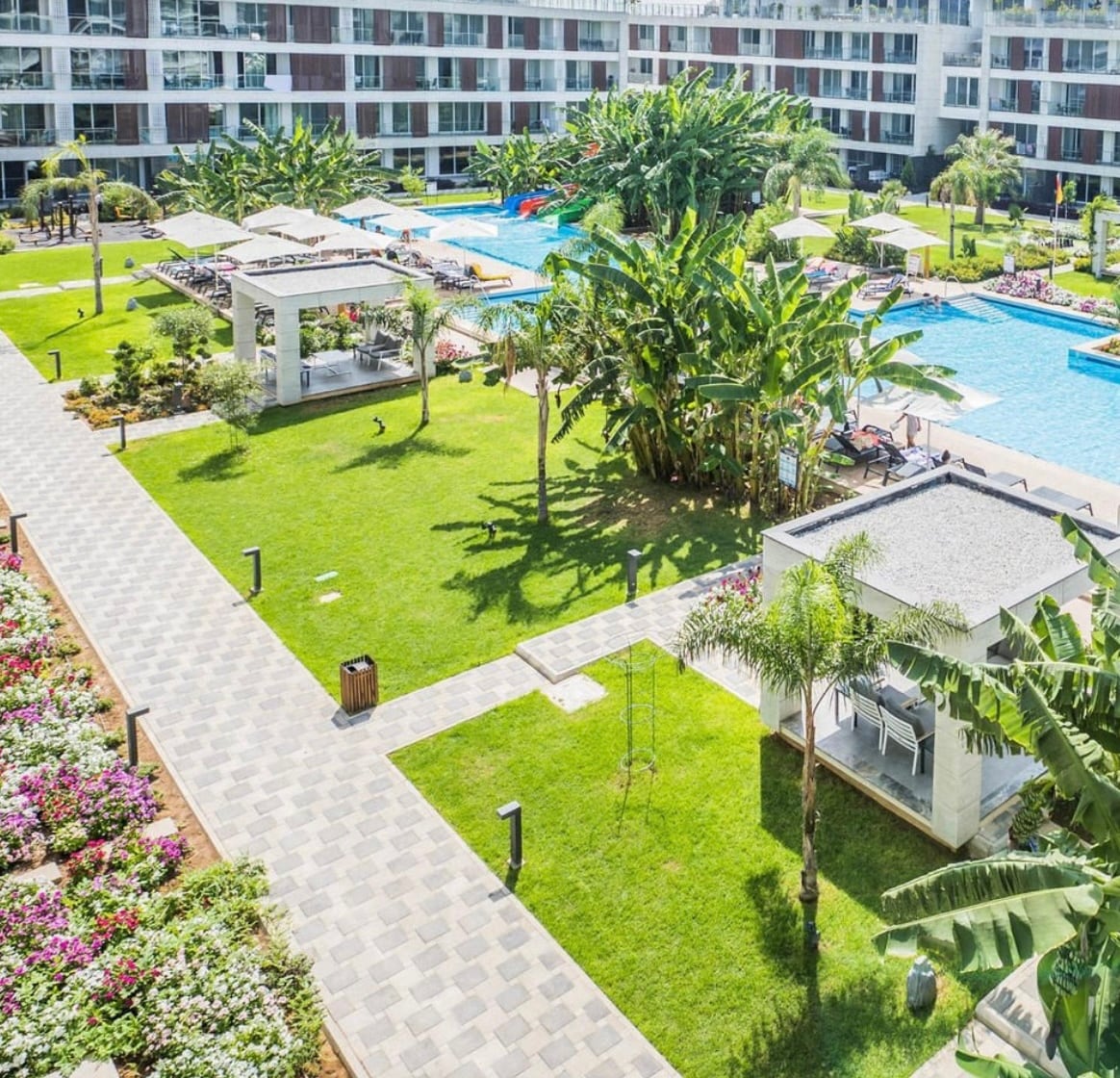 Aerial view of a lush courtyard featuring vibrant flower beds and manicured lawns. Sun loungers are positioned around a large pool, with shaded cabanas and palm trees enhancing the landscape. Pathways of stone lead through the greenery, inviting relaxation and outdoor activities.