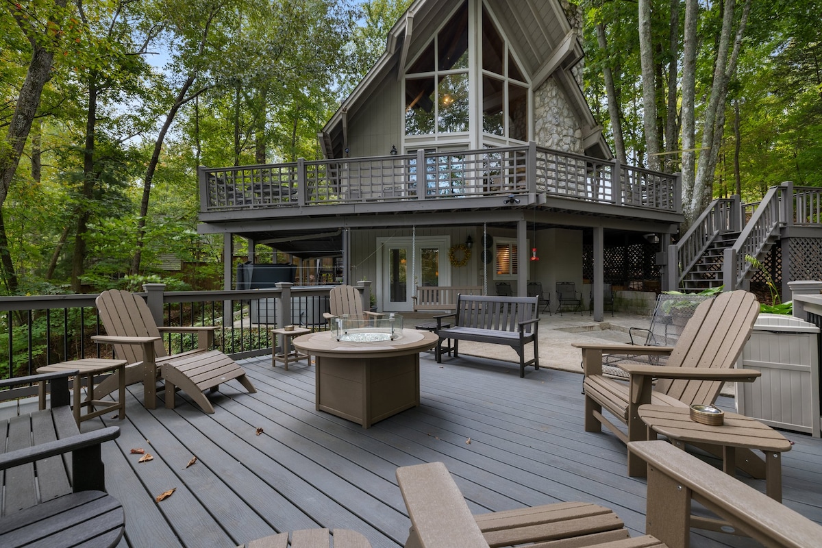 A deck area features multiple wooden lounge chairs arranged around a central fire pit. The cabin above has large windows that provide a view of the surrounding trees. The natural setting contributes to a serene outdoor space perfect for gatherings.