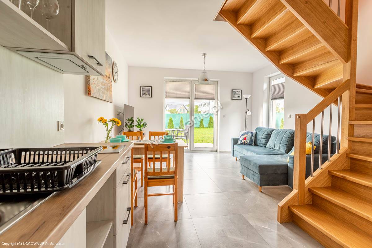 An open-concept living area features a dining table with four chairs, a fully equipped kitchen, and a comfortable seating arrangement. Natural light enters through large windows, illuminating the space. A staircase with wooden steps leads to the upper level, enhancing the inviting atmosphere.