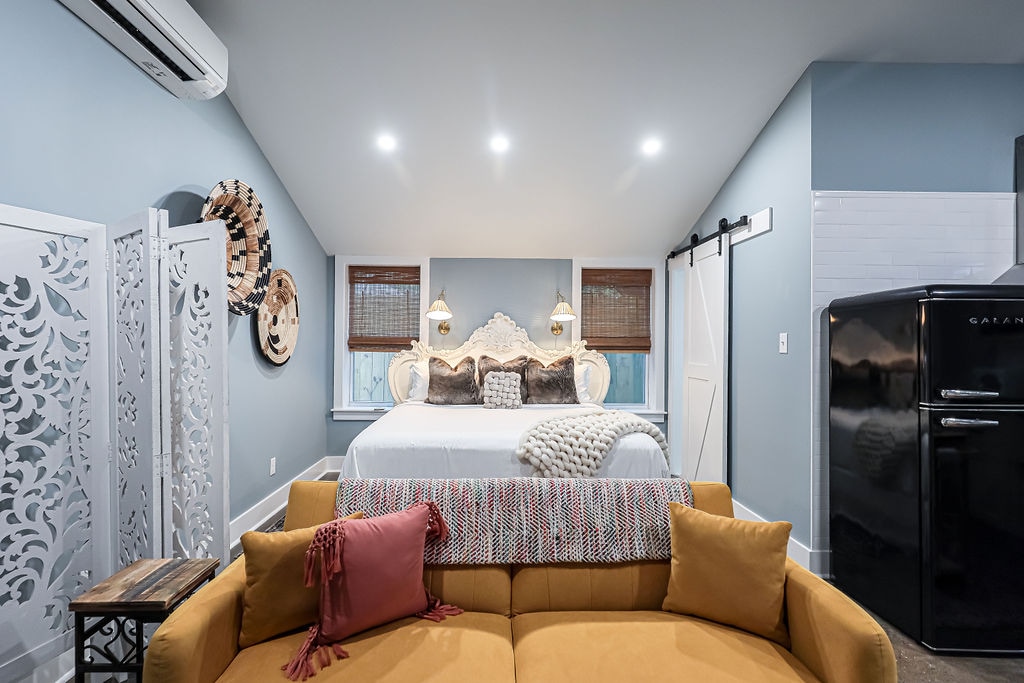 A cozy studio space is presented, featuring a spacious king bed with decorative pillows and soft linens. Flanking the bed are stylish lamps providing ambient light. A modern sofa in warm tones is positioned in the foreground, while a retro black refrigerator is visible in the corner.