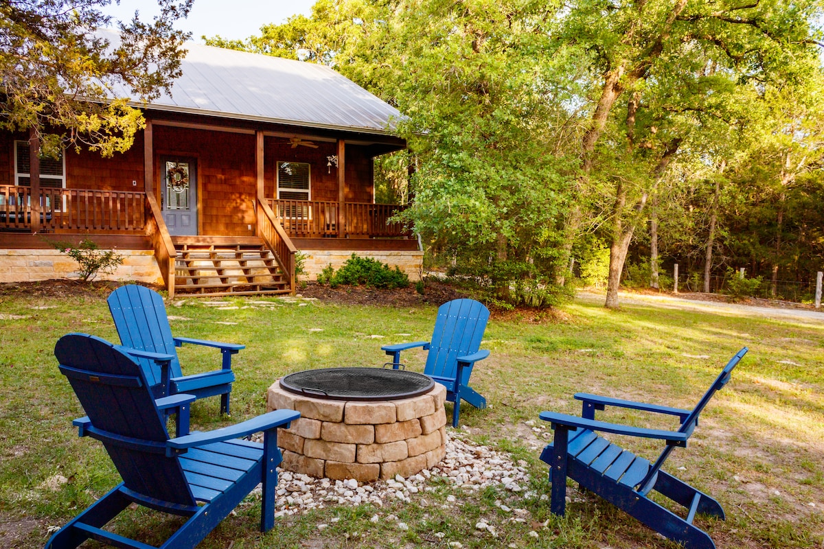 A charming cottage framed by lush greenery, featuring a circular fire pit surrounded by four blue Adirondack chairs. The entrance steps lead to a welcoming porch, inviting relaxation in a serene outdoor setting.